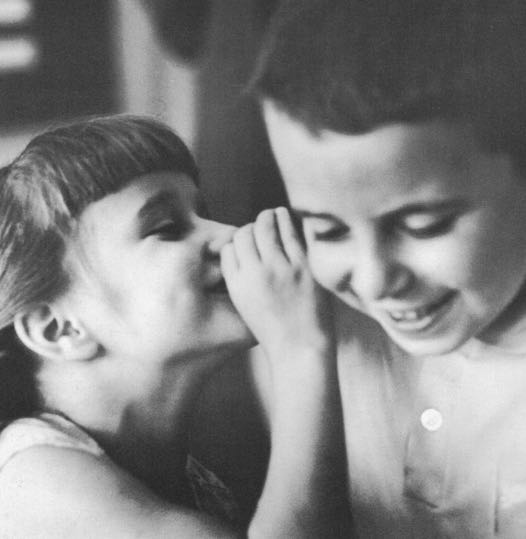 A young girl whispers something into the ear of a young boy. Both children are smiling. This image appeared in Creative Playthings catalog.