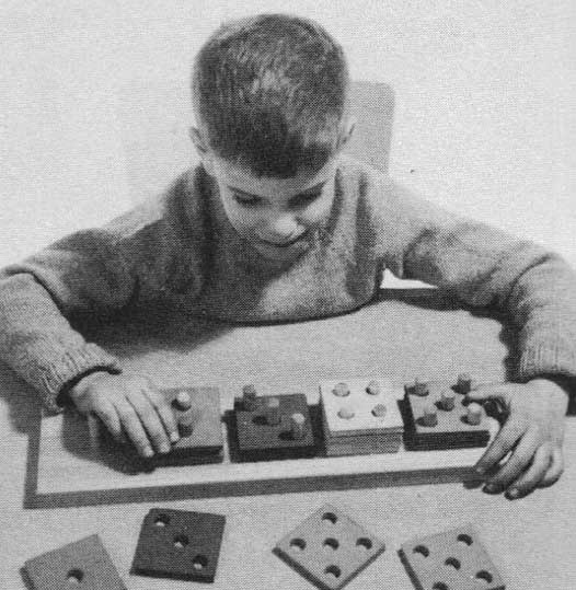 A young boy stacks squares with different arrangements of holes onto matching pegs. This image appeared in Creative Playthings catalog.